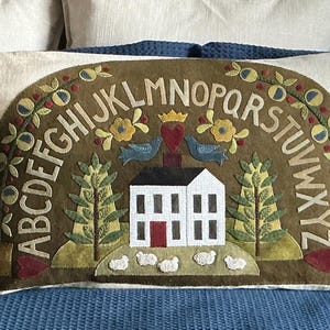 May include: A brown and green pillow with a white house, two birds, and a heart in the center. The alphabet is embroidered around the outside of the pillow.