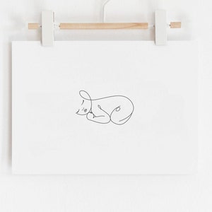 Puede incluir: Una impresi&oacute;n enmarcada en blanco con un dibujo de l&iacute;nea negra de un gato durmiendo. La impresi&oacute;n est&aacute; colgada en una pared blanca con una barra de madera y clips blancos.