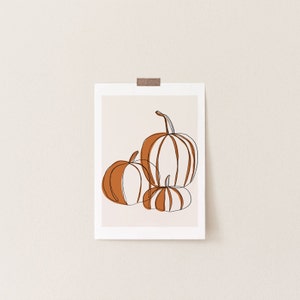 A seasonal fall print. This fall print has three pumpkins draw with a continuous black line. Random parts of the three pumpkins are painted orange and the background is a beige with a white border.