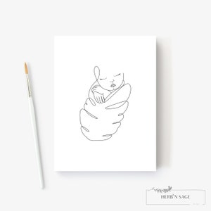 May include: A minimalist line art print of a sleeping baby swaddled in a leaf-like shape. The artwork is in black against a white background. A paintbrush is in the image. The print has the text "HERB'N SAGE design" at the bottom.