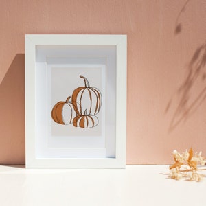 A seasonal fall print. This fall print has three pumpkins draw with a continuous black line. Random parts of the three pumpkins are painted orange and the background is a beige with a white border.