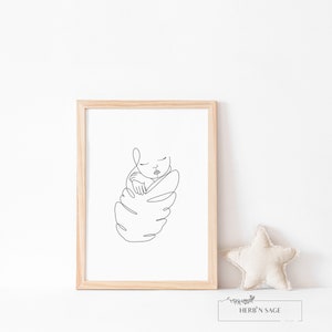 May include: A framed minimalist line art print of a sleeping baby. The artwork is in black on a white background, encased in a light wood frame. A beige star-shaped pillow and a small sign with the text "HERB'N SAGE" are next to the frame.