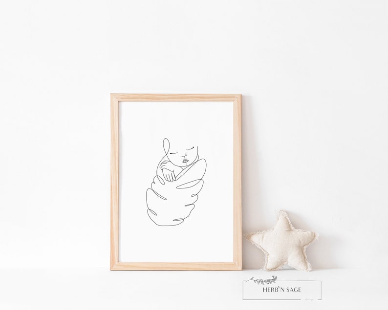 May include: A framed minimalist line art print of a sleeping baby. The artwork is in black on a white background, encased in a light wood frame. A beige star-shaped pillow and a small sign with the text "HERB'N SAGE" are next to the frame.