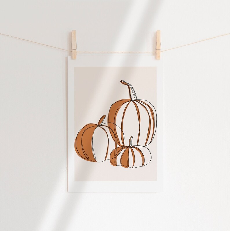 A seasonal fall print. This fall print has three pumpkins draw with a continuous black line. Random parts of the three pumpkins are painted orange and the background is a beige with a white border.