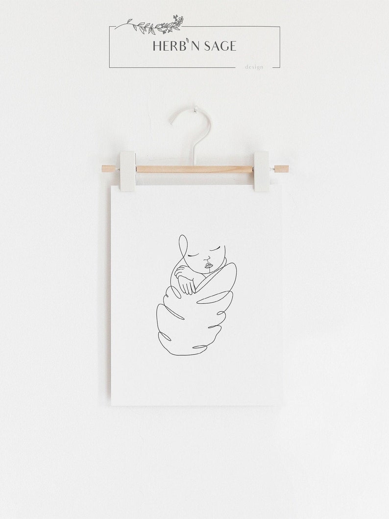 May include: A minimalist line art print of a baby swaddled in a leaf-like shape. The artwork is in black against a white background, displayed on a wooden hanger. The print includes the text "HERB'N SAGE design" at the top.
