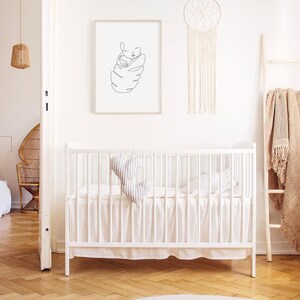 May include: A minimalist nursery scene with a white crib, a framed line art print of a baby, and a macrame dream catcher. A wooden ladder holds a beige blanket. The room has a neutral color palette with wooden floors.
