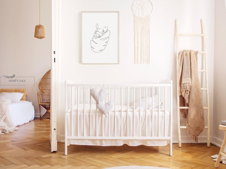 May include: A minimalist nursery scene with a white crib, a framed line art print of a baby, and a macrame dream catcher. A wooden ladder holds a beige blanket. The room has a neutral color palette with wooden floors.