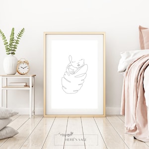 May include: A framed minimalist line art print featuring a sleeping baby. The black line drawing is set against a white background within a gold frame. The print is displayed on a light wood floor, with a white vase and clock visible.