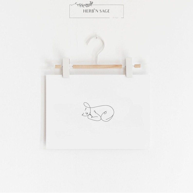 Puede incluir: Una impresi&oacute;n enmarcada en blanco con un dibujo de l&iacute;nea negra de un gato durmiendo. La impresi&oacute;n est&aacute; colgada en una pared blanca con una barra de madera y clips blancos.