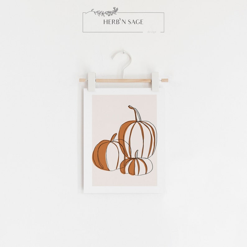 A seasonal fall print. This fall print has three pumpkins draw with a continuous black line. Random parts of the three pumpkins are painted orange and the background is a beige with a white border.