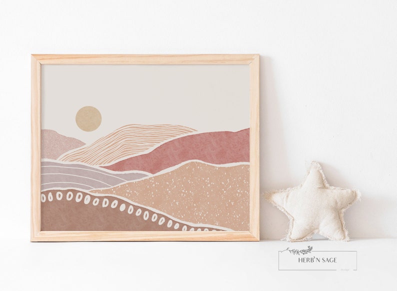Peut inclure: Une impression encadr&eacute;e d'un paysage minimaliste avec un soleil, des collines et un fond textur&eacute; dans des tons de brun, rose et beige. L'impression est dans un cadre en bois clair.