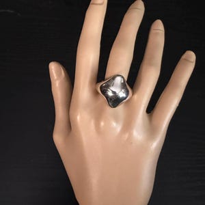 May include: A silver ring with an irregular, organic shape. The ring is highly polished and reflects light, creating a bright, shiny appearance. The ring is shown on a mannequin hand against a dark background. The ring is a statement piece.