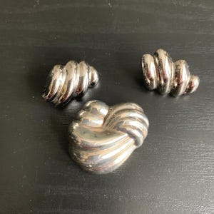 May include: A set of silver-toned jewelry, including a brooch and two matching earrings. The brooch is a knot design, and the earrings have a spiral shape. The jewelry is displayed on a dark surface.