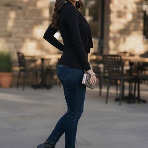 Puede incluir: Una persona con una blusa negra de manga larga, vaqueros azul oscuro y botines negros de tacón. Sostiene un pequeño bolso de mano plateado. El fondo incluye un edificio de ladrillo y asientos al aire libre.