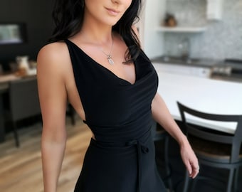 May include: A black dress with a cowl neckline and a tie waist. The dress has a sleeveless design and a short skirt. The model is wearing a silver necklace and bracelet.