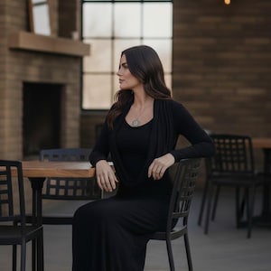 May include: A woman wearing a black long-sleeve dress with a draped neckline, seated at a wooden table. She is wearing a silver pendant necklace. The setting is outdoors, with a brick wall and large window in the background.