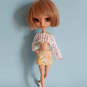 Op de afbeelding: Een pop met blond haar die een wit gehaakt topje, een rok met bloemenprint en witte sneakers draagt.