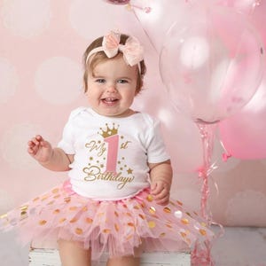 May include: A baby girl wearing a white onesie with a pink and gold "My 1st Birthday" graphic, a pink tulle tutu with gold polka dots, and a pink and gold headband. The baby is sitting in front of a pink background with balloons.