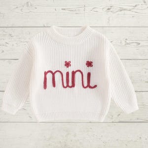 May include: White knitted sweater with long sleeves and a crew neck. The sweater has the word "mini" embroidered in red on the front.
