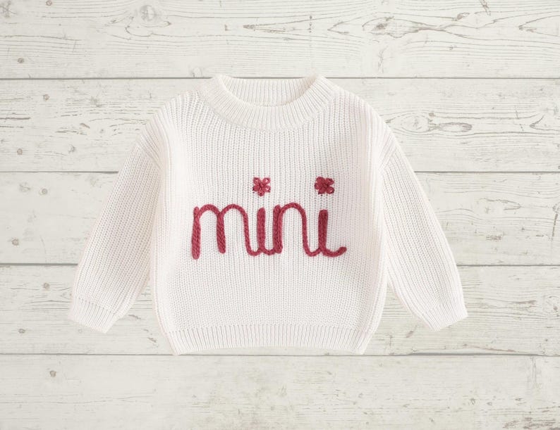 May include: White knitted sweater with long sleeves and a crew neck. The sweater has the word "mini" embroidered in red on the front.