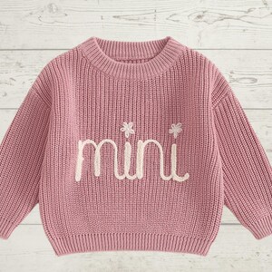 May include: A pink knitted sweater with long sleeves and a crew neck. The sweater has the word "mini" embroidered on the front in white thread.