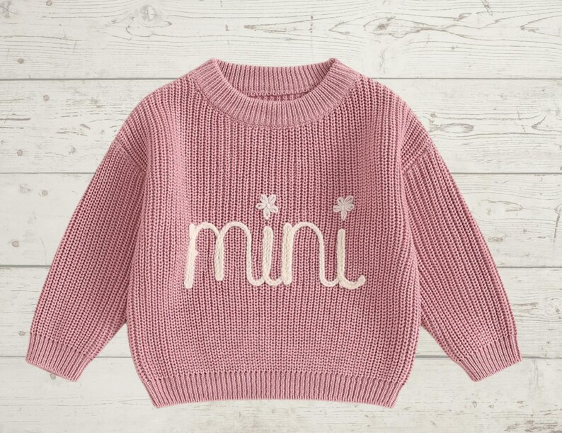 May include: A pink knitted sweater with long sleeves and a crew neck. The sweater has the word "mini" embroidered on the front in white thread.