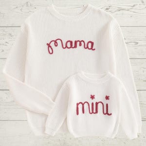 May include: White knitted sweaters with red embroidered text. The larger sweater says "mama" and the smaller sweater says "mini".