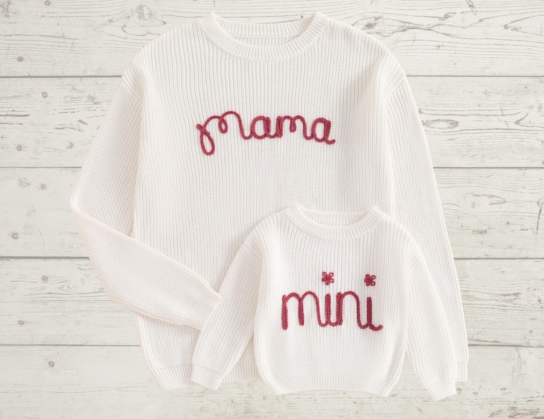 May include: White knitted sweaters with red embroidered text. The larger sweater says "mama" and the smaller sweater says "mini".