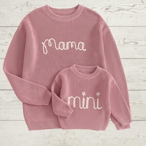 May include: A pink knitted sweater set with the word "mama" embroidered on the larger sweater and the word "mini" embroidered on the smaller sweater.