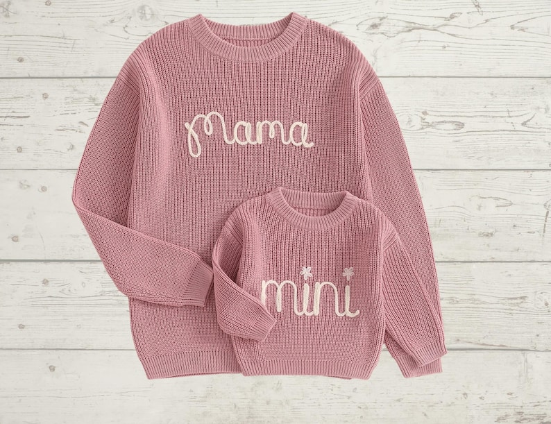May include: A pink knitted sweater set with the word "mama" embroidered on the larger sweater and the word "mini" embroidered on the smaller sweater.