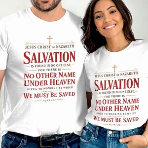 Peut inclure: Deux t-shirts blancs à manches courtes avec une croix dorée et un texte rouge indiquant : Jesus Christ of Nazareth, Salvation is found in no one else, for there is no other name under heaven given to mankind by which we must be saved, Acts 4:12.