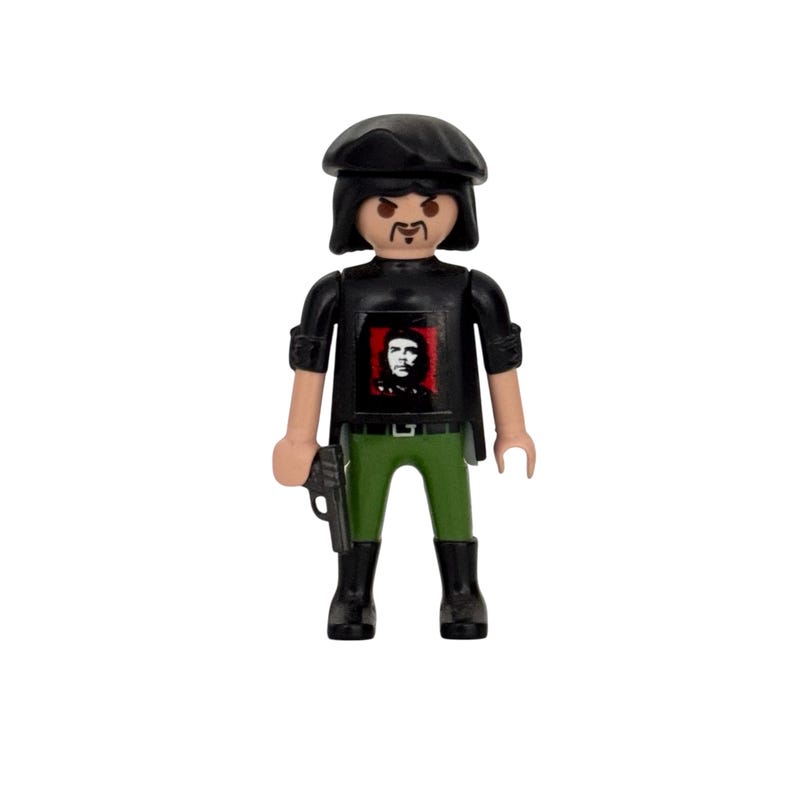 May include: A black and green plastic figurine of a person wearing a black beret, black shirt with a red and black image of Che Guevara, and green pants. The figurine is holding a black gun in its right hand.