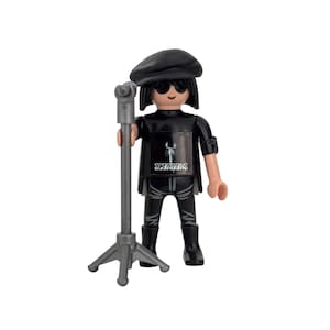 Puede incluir: Figura negra y gris de una persona que lleva un gorro negro, gafas de sol y una camiseta negra con la palabra "SCORPIONS" y un gráfico de escorpión. La figura sostiene un soporte de micrófono.