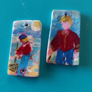 Peut inclure: Deux pendentifs rectangulaires en céramique avec des scènes peintes à la main. Le pendentif de gauche montre une silhouette en chemise rouge et pantalon bleu près d'un soleil jaune. Celui de droite représente une silhouette en chemise rouge.