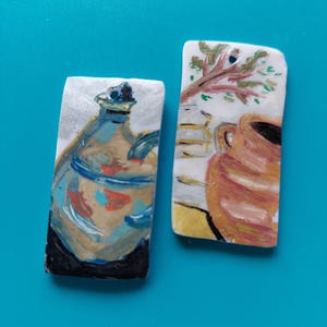 Peut inclure: Deux carreaux de céramique rectangulaires avec des images peintes. L'un représente une cruche bleue et beige avec une base sombre. L'autre montre un vase marron et une branche d'arbre avec des feuilles vertes. Le fond est blanc.