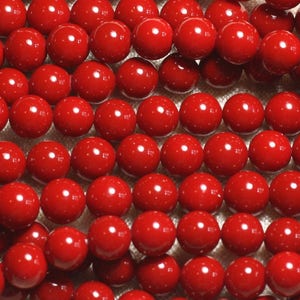 May include: Close-up of a collection of bright red, round beads. The beads are evenly spaced and appear to be strung together, possibly for jewelry making. The beads have a glossy finish and reflect light, creating a vibrant and eye-catching display.
