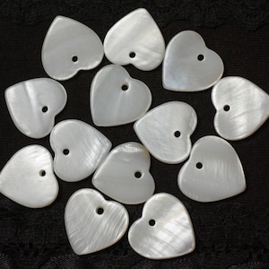 Peut inclure: Un ensemble de 14 boutons en forme de cœur en coquille blanche avec un trou au centre. Les boutons sont disposés de manière aléatoire sur un fond en tissu noir.