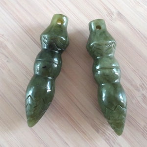 Può includere: Due ciondoli in giada verde con dettagli scolpiti. Ogni ciondolo ha una forma cilindrica con un foro nella parte superiore per l'infilatura. I ciondoli sono di colore verde scuro e hanno una finitura liscia e lucida.