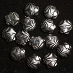 Pode incluir: Uma coleção de 15 pingentes de concha do mar de metal prateado. Os pingentes têm todos a forma de uma concha de vieira com um design em relevo detalhado.