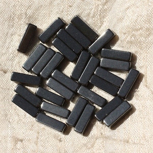 Peut inclure: Un groupe de perles rectangulaires noires, probablement en hématite, éparpillées sur un fond de tissu blanc.