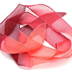 May include: A length of sheer, red ribbon, folded and layered against a white background. The ribbon's color transitions from a deep burgundy to a vibrant coral hue. The ribbon's edges are neatly finished.