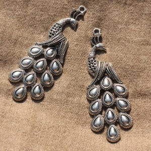 May include: Two silver peacock charms with detailed designs. Each charm features a peacock with a spread tail of teardrop-shaped accents.