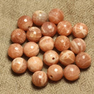 May include: A collection of round, peach-colored sunstone beads with faceted surfaces. The beads have natural variations in color and texture, and are arranged on a textured, tan surface. These beads are ideal for jewelry making.