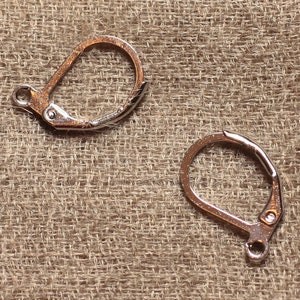 May include: Two silver-toned earring hooks, also known as ear wires, are shown on a brown textured surface. The hooks are made of metal and have a simple design.