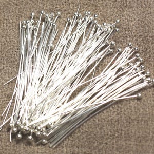 Op de afbeelding: Een grote stapel zilverkleurige, rechte, dunne metalen pinnen met ronde koppen. De pinnen zijn in een willekeurig patroon gerangschikt en liggen op een bruin stofoppervlak.