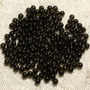 May include: A pile of small, round, black beads. The beads are scattered on a textured, off-white surface. The beads appear to be made of a shiny, reflective material.