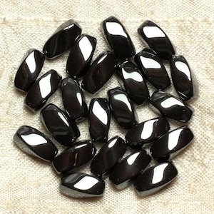 May include: A collection of polished, black gemstone beads. Each bead is oval-shaped with a smooth, reflective surface. The beads are scattered on a textured, light-colored fabric, creating a contrast between the dark stones and the neutral background.