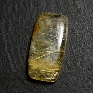 Può includere: Un cabochon di quarzo trasparente con inclusioni di rutilo dorate. La pietra è di colore giallo chiaro e ha una superficie liscia e lucida.