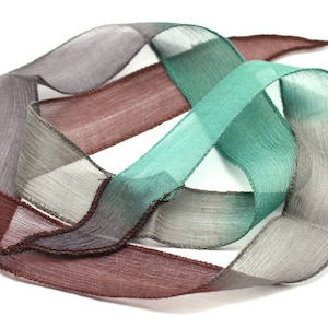 May include: A close-up of a brown, gray, and teal ribbon with a textured, woven look. The ribbon is folded and twisted into a decorative shape.