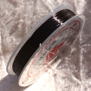 Pode incluir: Um carretel de fio de cristal preto, enrolado firmemente e embalado em um recipiente de plástico transparente. O carretel é rotulado com texto japonês e um gráfico vermelho. Para fazer joias ou artesanato.